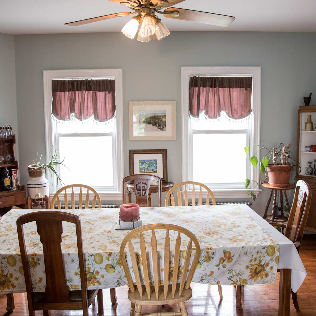 A dining room features a large table surrounded by wooden chairs, with windows adorned by roman shades and a sideboard displaying glassware.
