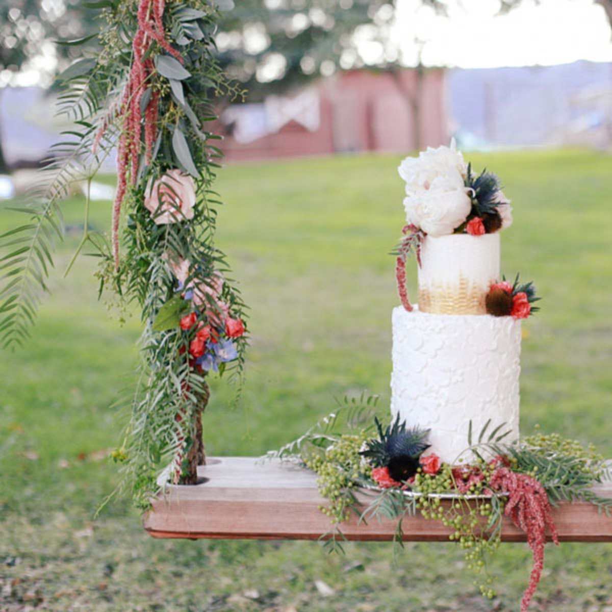 A two-tiered wedding cake adorned with flowers and greenery is displayed on a wooden swing in a grassy outdoor setting.