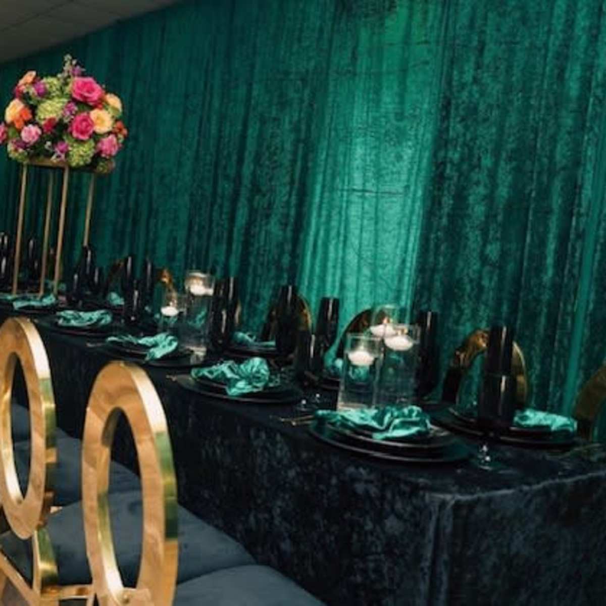A long dining table is set with black tableware and surrounded by gold chairs, decorated with colorful floral centerpieces against a green backdrop.
