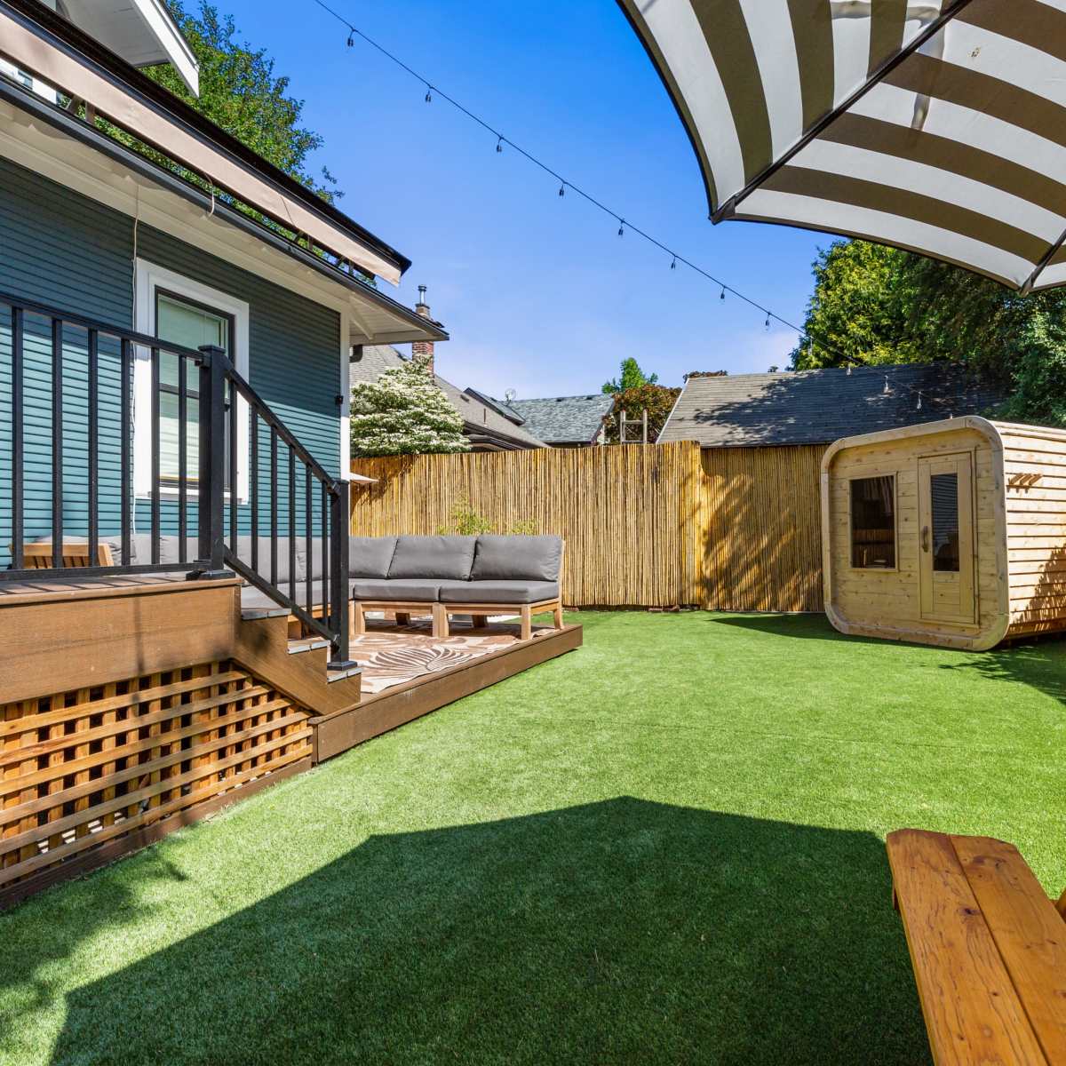 A backyard with a green artificial lawn, a wooden picnic table, a lounge area with gray seating, and a small wooden structure against a wooden fence.