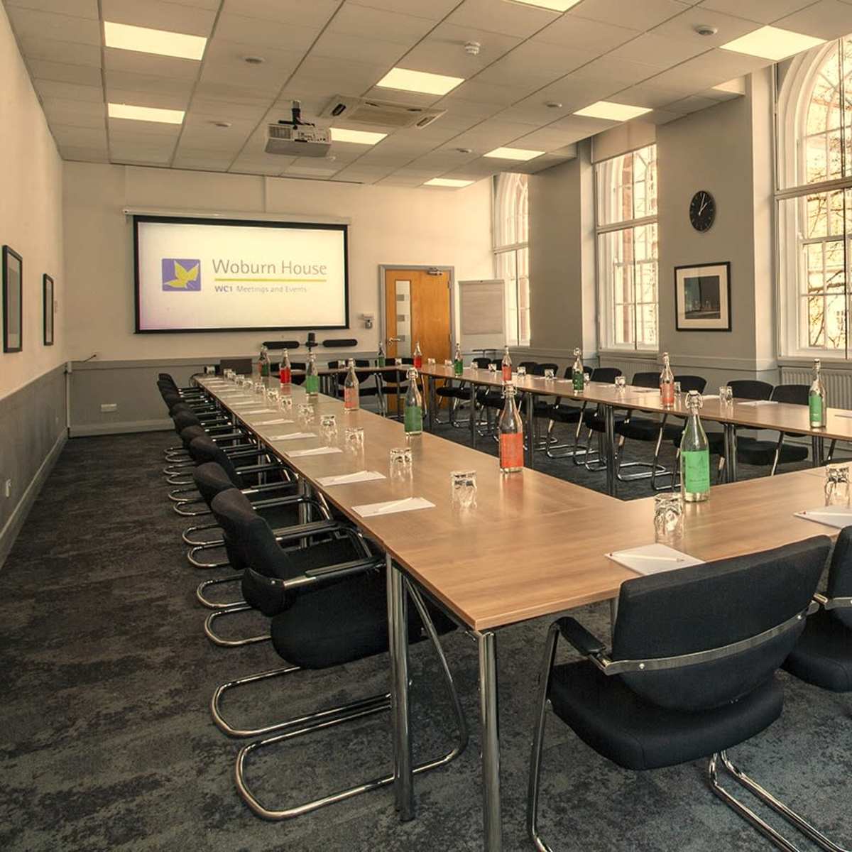 A meeting room is set up with rectangular tables arranged in a U-shape, featuring chairs and various drinks on the tables, with a presentation screen at one end.