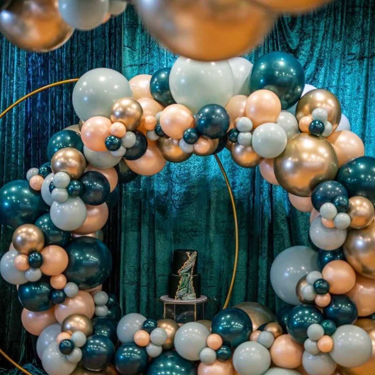 A decorative balloon arch in shades of teal, peach, and gold, set against a shimmering green backdrop.