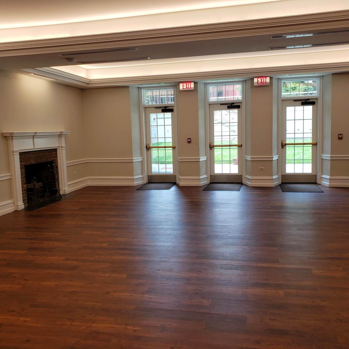 A spacious, empty room with hardwood floors, a fireplace on one side, and three windows with exit doors leading outside.