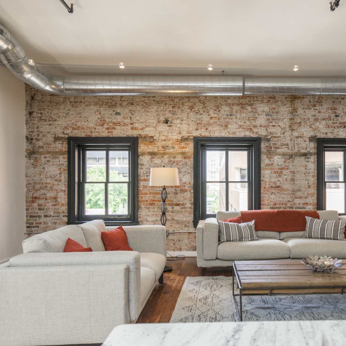 The room features a rustic brick wall with large windows, two light-colored sofas adorned with orange cushions, and a wooden coffee table.