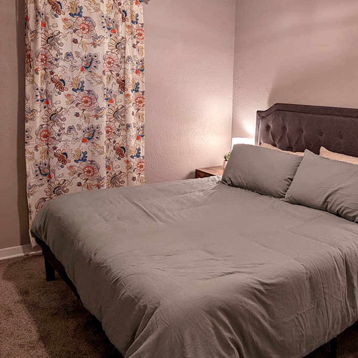 A neatly arranged bedroom with a gray bedspread, two pillows, and decorative curtains with floral patterns, beside wooden nightstands and lamps.