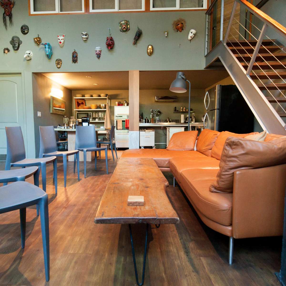 A living room with a brown leather sofa, a wooden coffee table, several gray chairs arranged around the room, and a staircase leading to an upper level adorned with various masks on the walls.