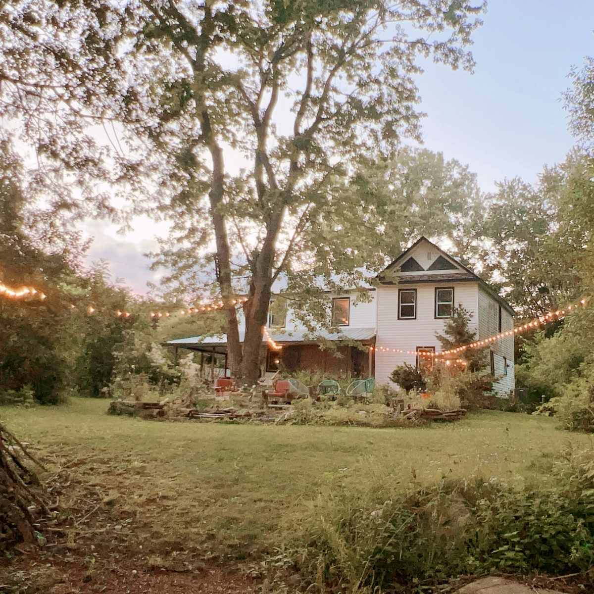 A house surrounded by trees and a well-kept garden is illuminated by string lights, creating a cozy atmosphere.