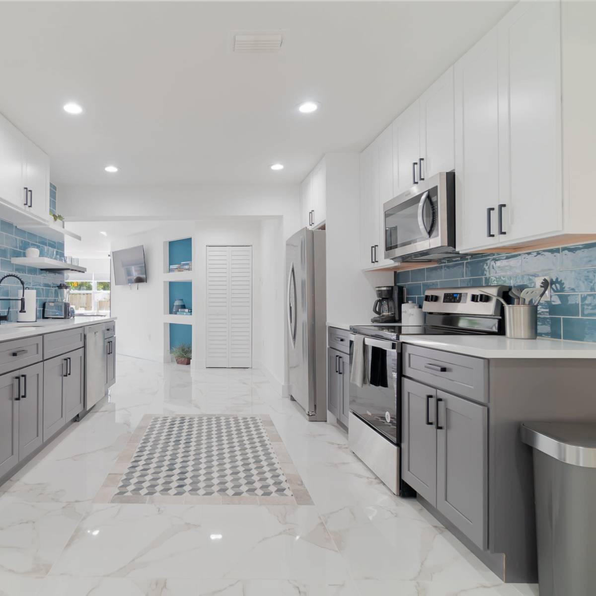 A modern kitchen featuring gray and white cabinetry, blue tiled accents, stainless steel appliances, and a marble-patterned floor.