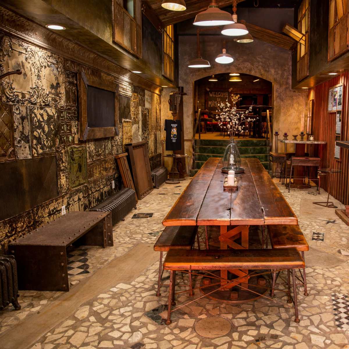 A rustic dining area with a long wooden table, stone flooring, and vintage decor, featuring walls with various textures and framed images.