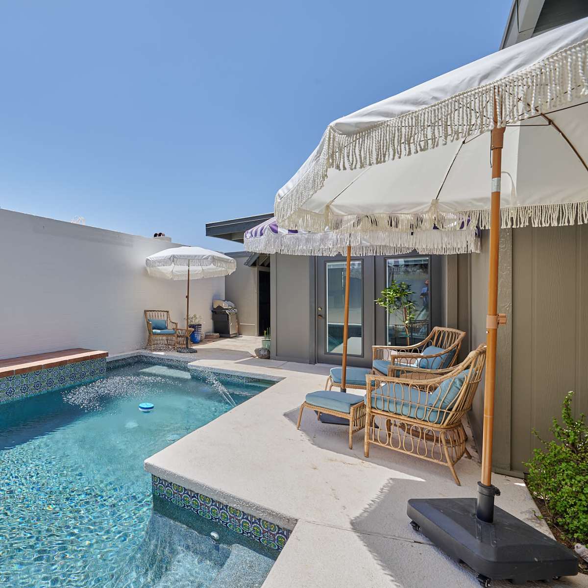 A small swimming pool is situated next to a shaded seating area with umbrellas in a walled courtyard.
