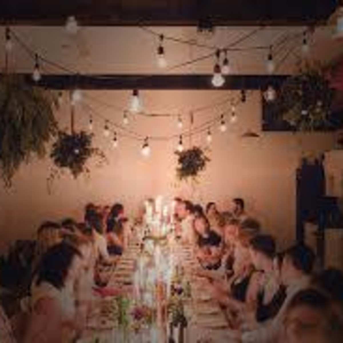 A long table is set for a dinner party, lined with candles and surrounded by guests in a warmly lit space with hanging plants and string lights overhead.