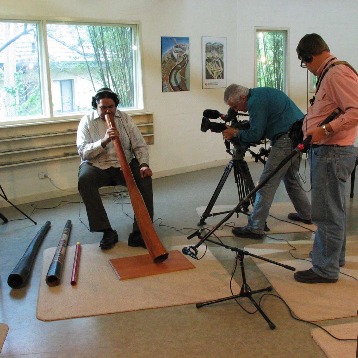 A man plays a didgeridoo while two cameramen record the performance in a room with large windows and colorful artwork.