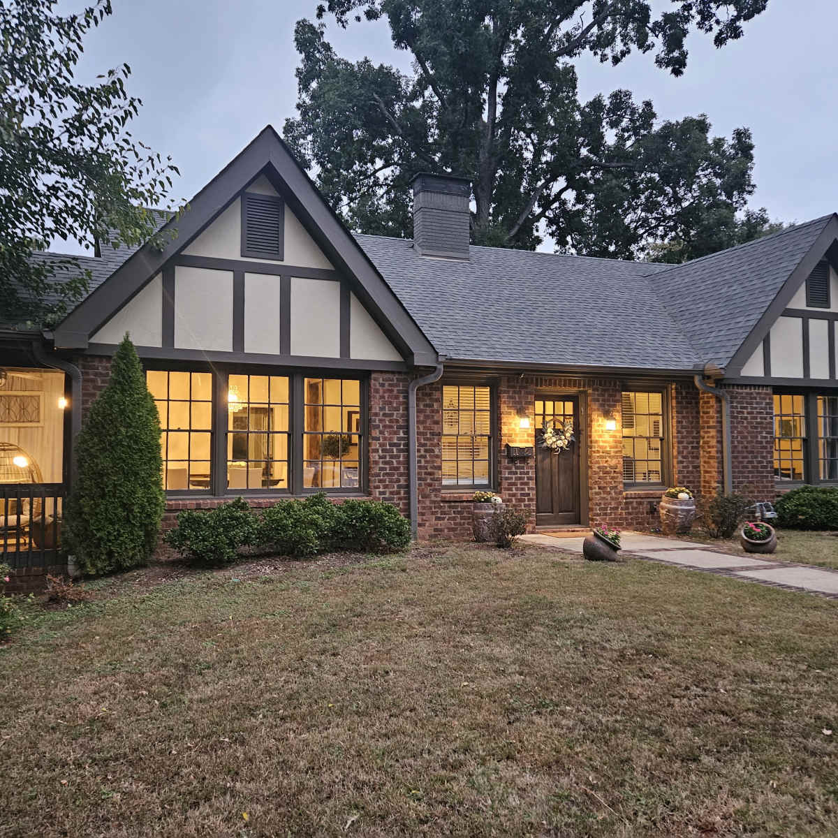 A Tudor-style brick house with a steeply pitched roof and large windows, surrounded by greenery and a neatly maintained lawn.