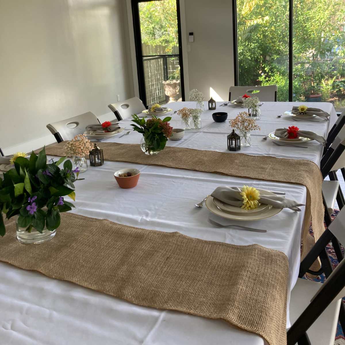 A dining room features two tables set with white tablecloths, decorated with small floral arrangements and simple dishware, overlooking a view of greenery through large windows.