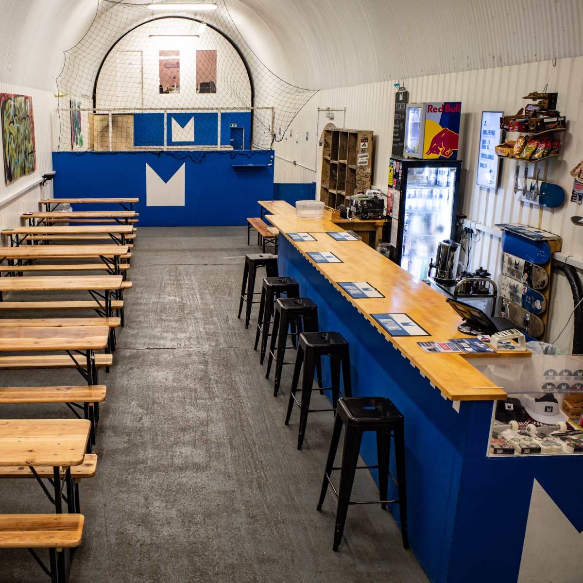 A spacious indoor venue with wooden tables, black stools, and a blue bar area, featuring graffiti on the walls and a netted section in the background.