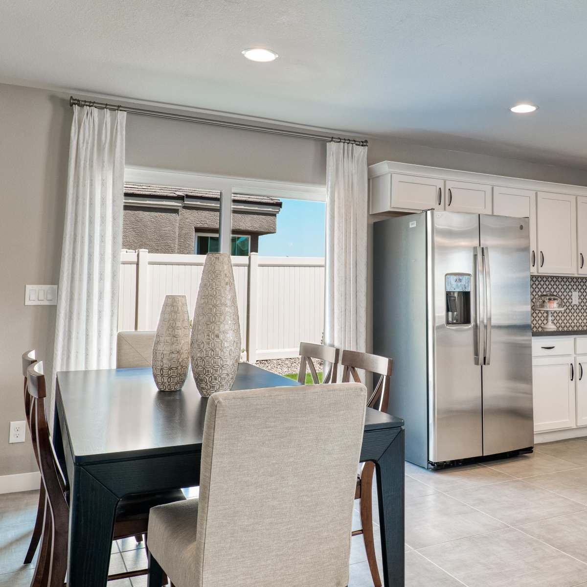 A modern kitchen features stainless steel appliances, a dining table, and large windows with sheer curtains.