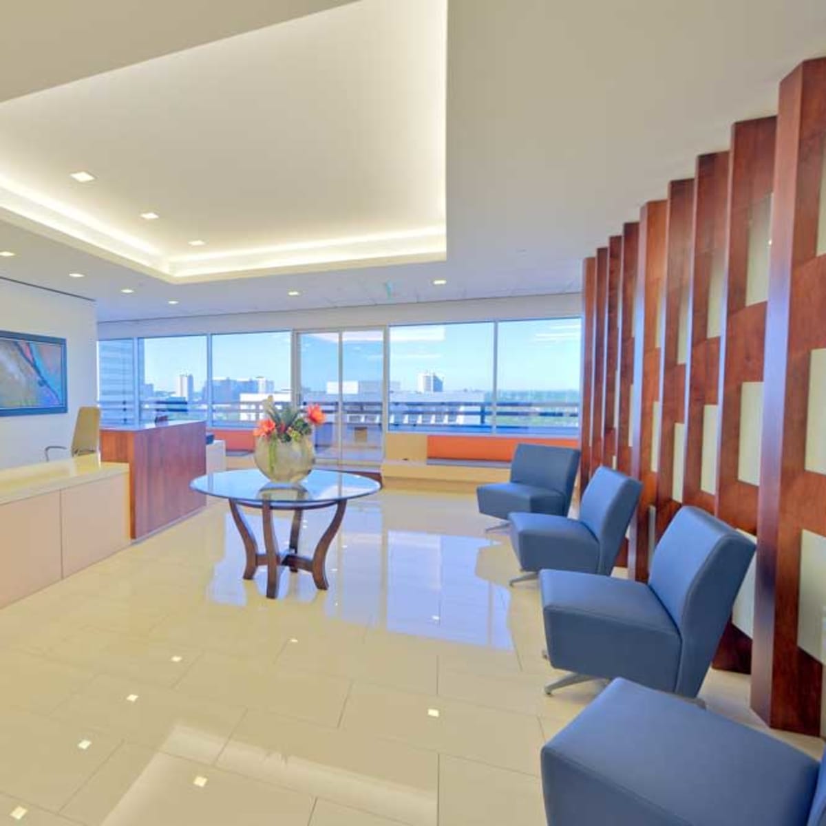 A modern office reception area featuring a sleek front desk, a round glass table with a decorative vase, blue chairs, and large windows with a city view.