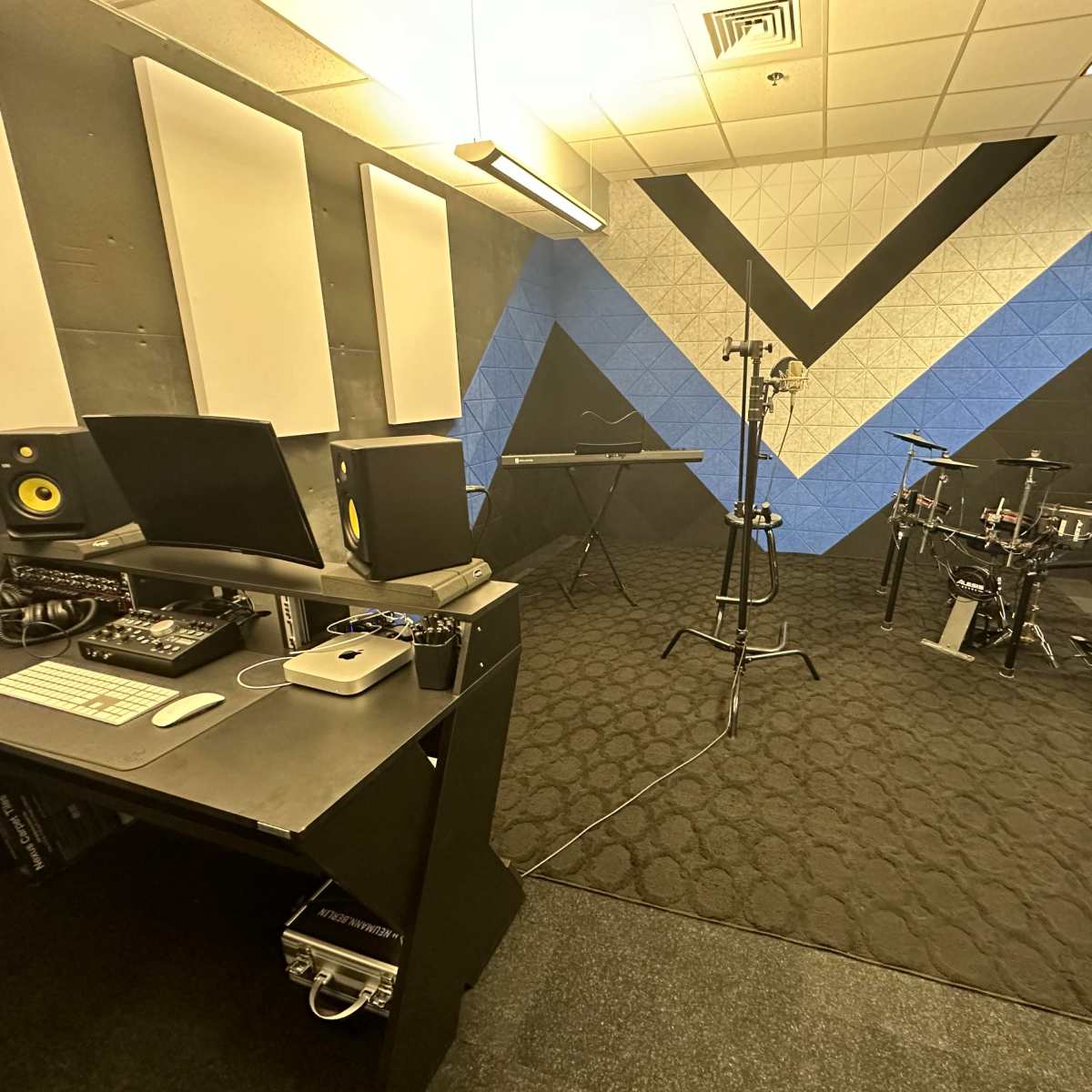 A music production room featuring a computer setup, an electronic keyboard, and a drum kit against a blue and black geometric wall.