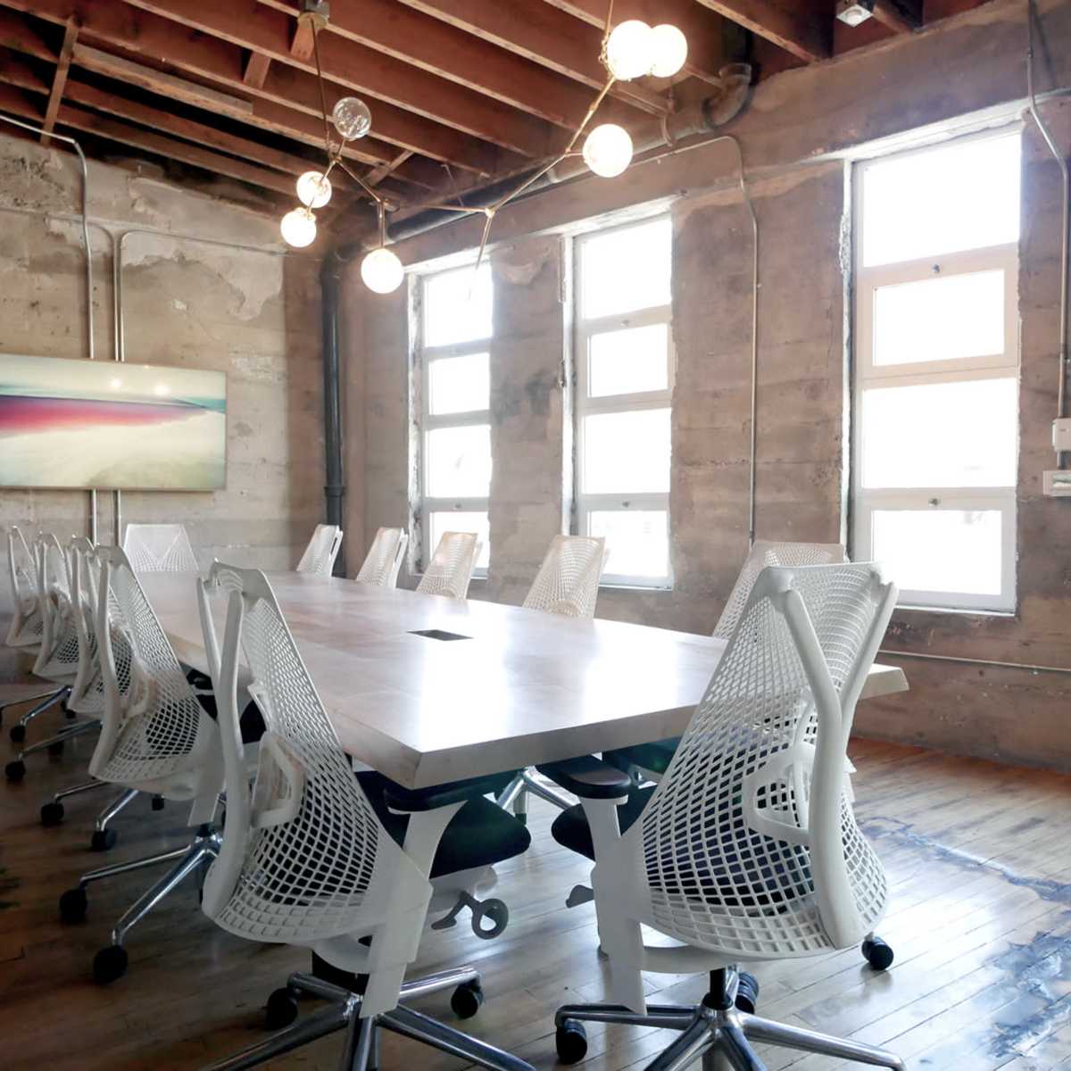 A long conference table with white mesh chairs is set up in a brightly lit room featuring exposed wooden beams and concrete walls.
