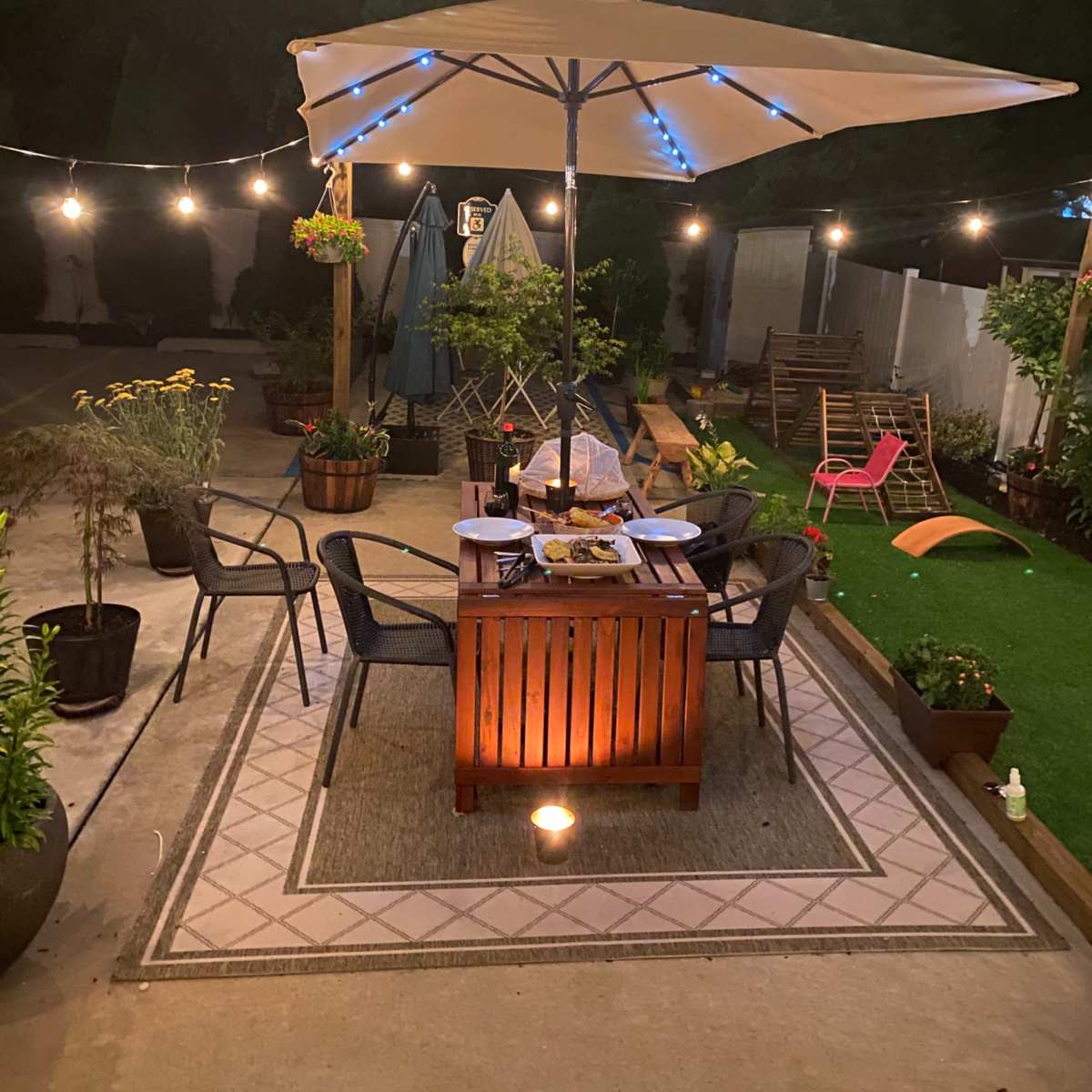 A patio area is illuminated by string lights and features a central table with food under a large umbrella, surrounded by chairs and various potted plants.