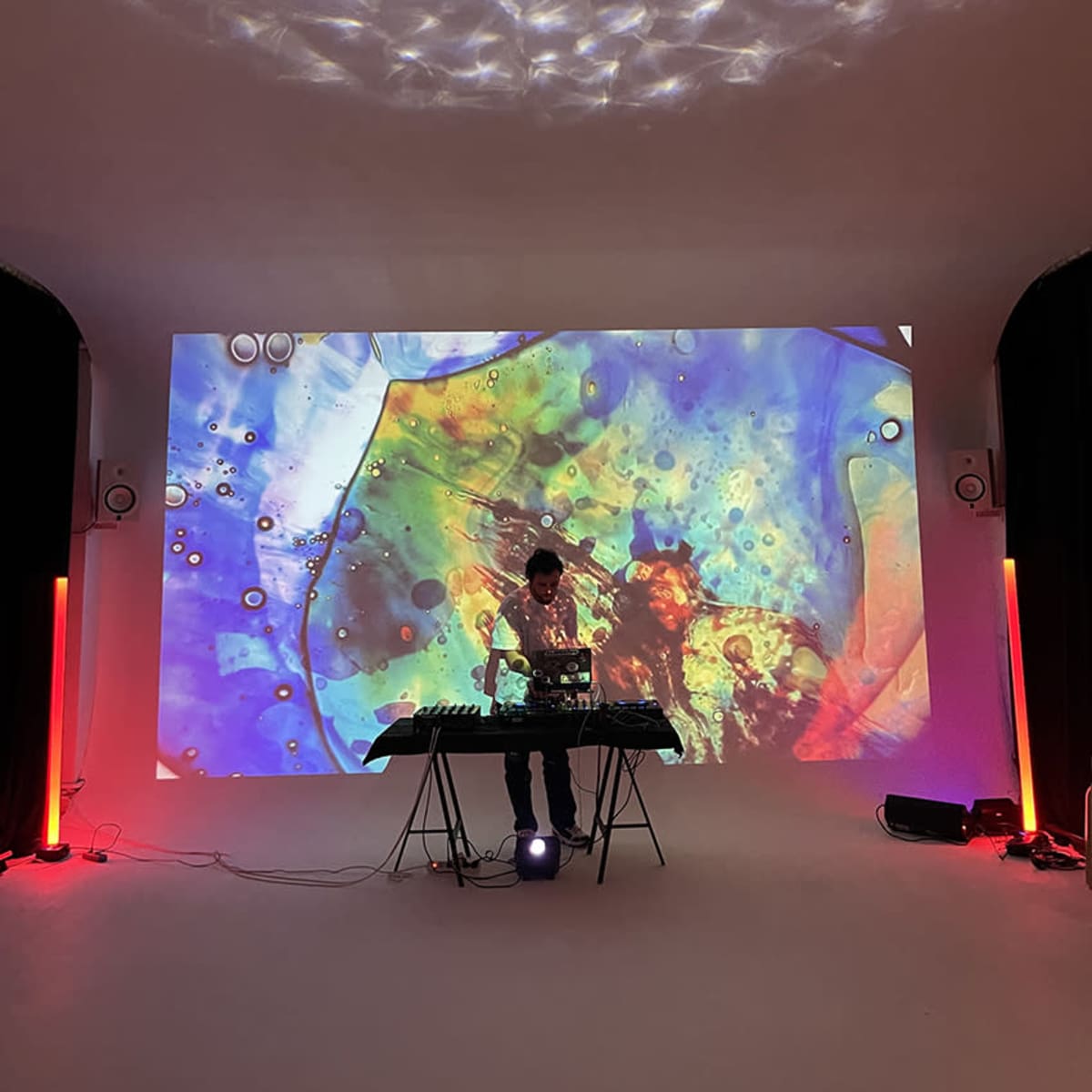 A person stands behind a music equipment setup in a room with colorful abstract visuals projected on a screen behind them.