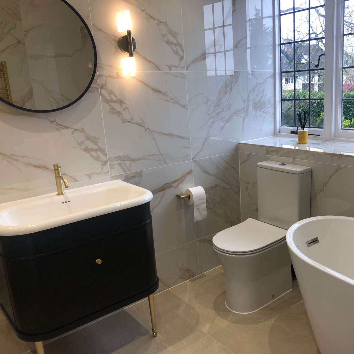 A modern bathroom featuring a freestanding bathtub, a toilet, and a vanity with a round mirror, all accented by marble-patterned walls and a window allowing natural light.