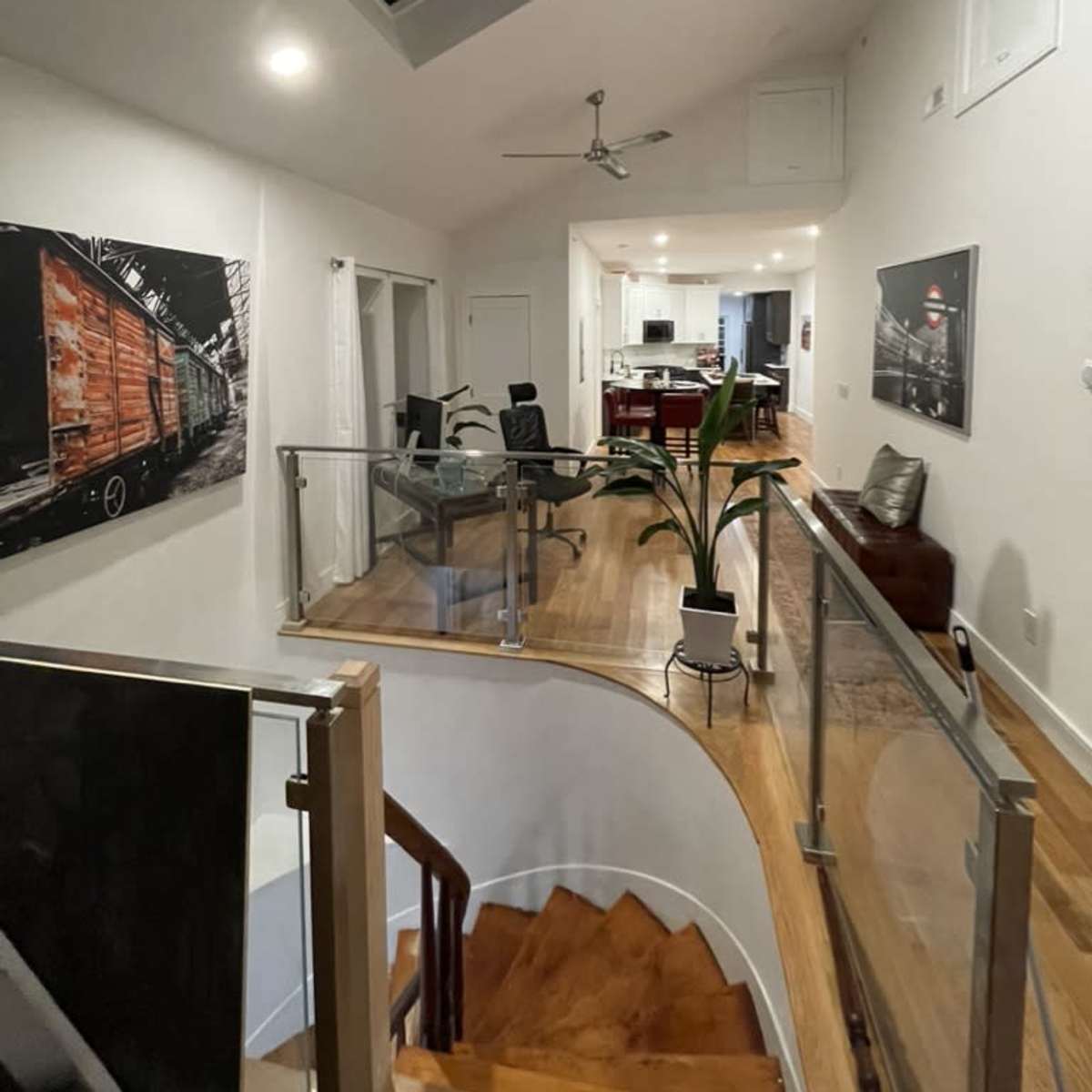 A modern hallway with a spiraling staircase, featuring artworks on the walls and a view into a kitchen and dining area in the background.