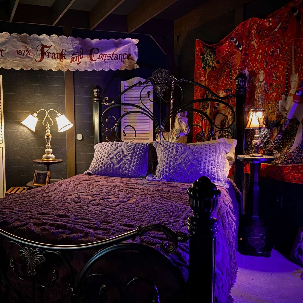 A dimly lit bedroom featuring a wrought iron bed with a purple bedspread, decorative pillows, and a patterned wall hanging.