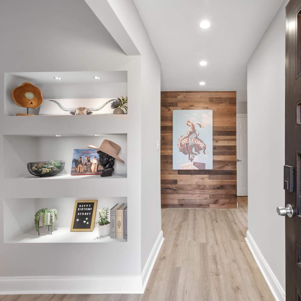 A hallway features a modern entry door, built-in shelves with decorative items, and a wood-paneled accent wall displaying artwork.