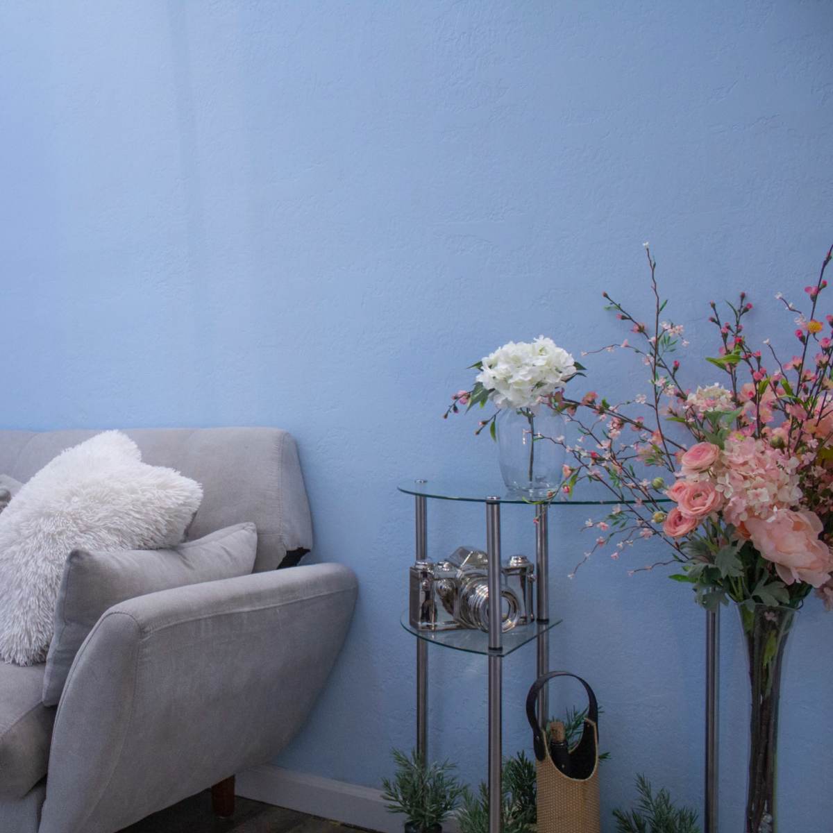 A light blue room featuring a gray sofa with pillows, a glass side table, and a vase of flowers.