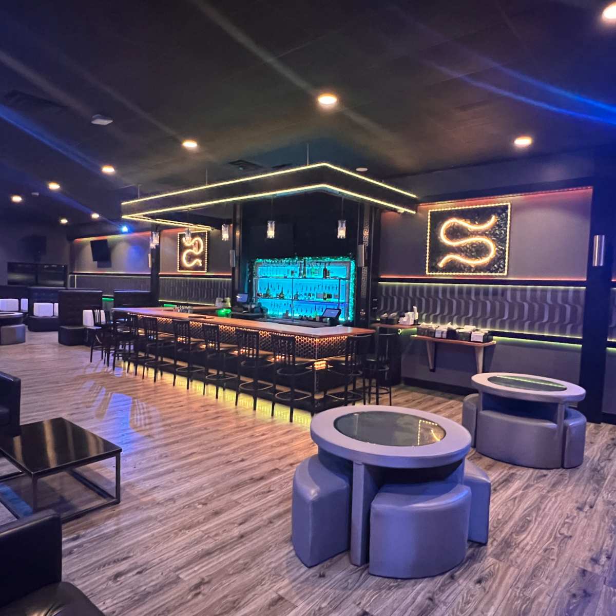A modern lounge interior with a sleek bar, illuminated by blue lighting, surrounded by black leather seating and circular tables.