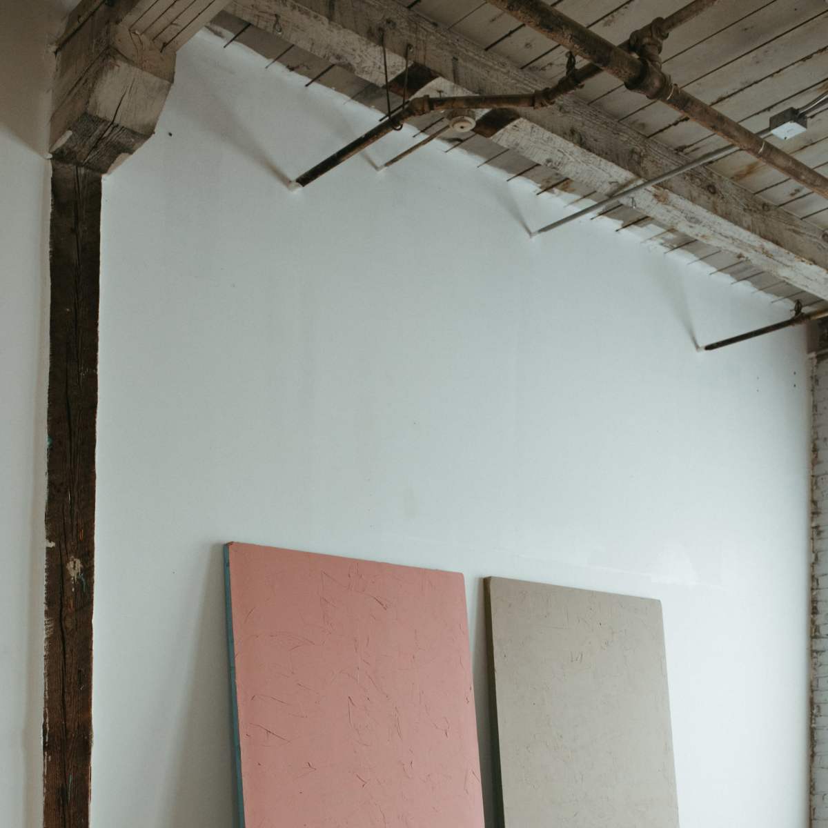 Two large, unframed panels, one pink and one beige, are leaning against a white wall in a room with wooden floors and exposed beams.