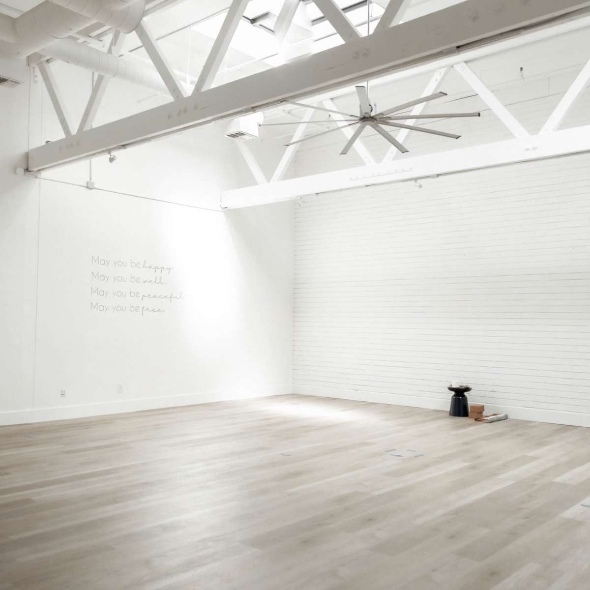 A spacious, minimalist room with white walls, wooden flooring, and a large ceiling fan.