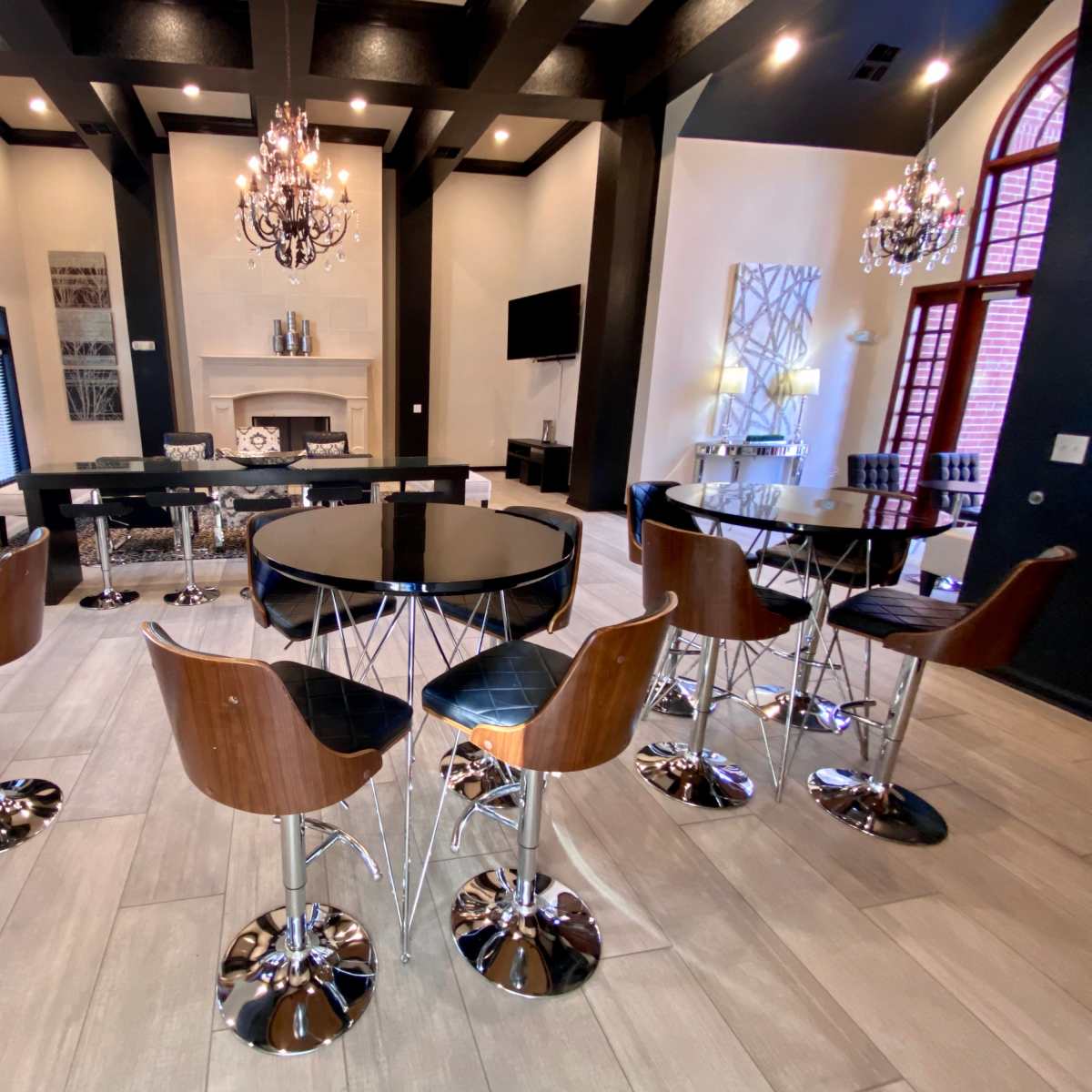 A modern lounge area with high bar stools arranged around circular tables, illuminated by chandeliers and featuring a fireplace and a large television.