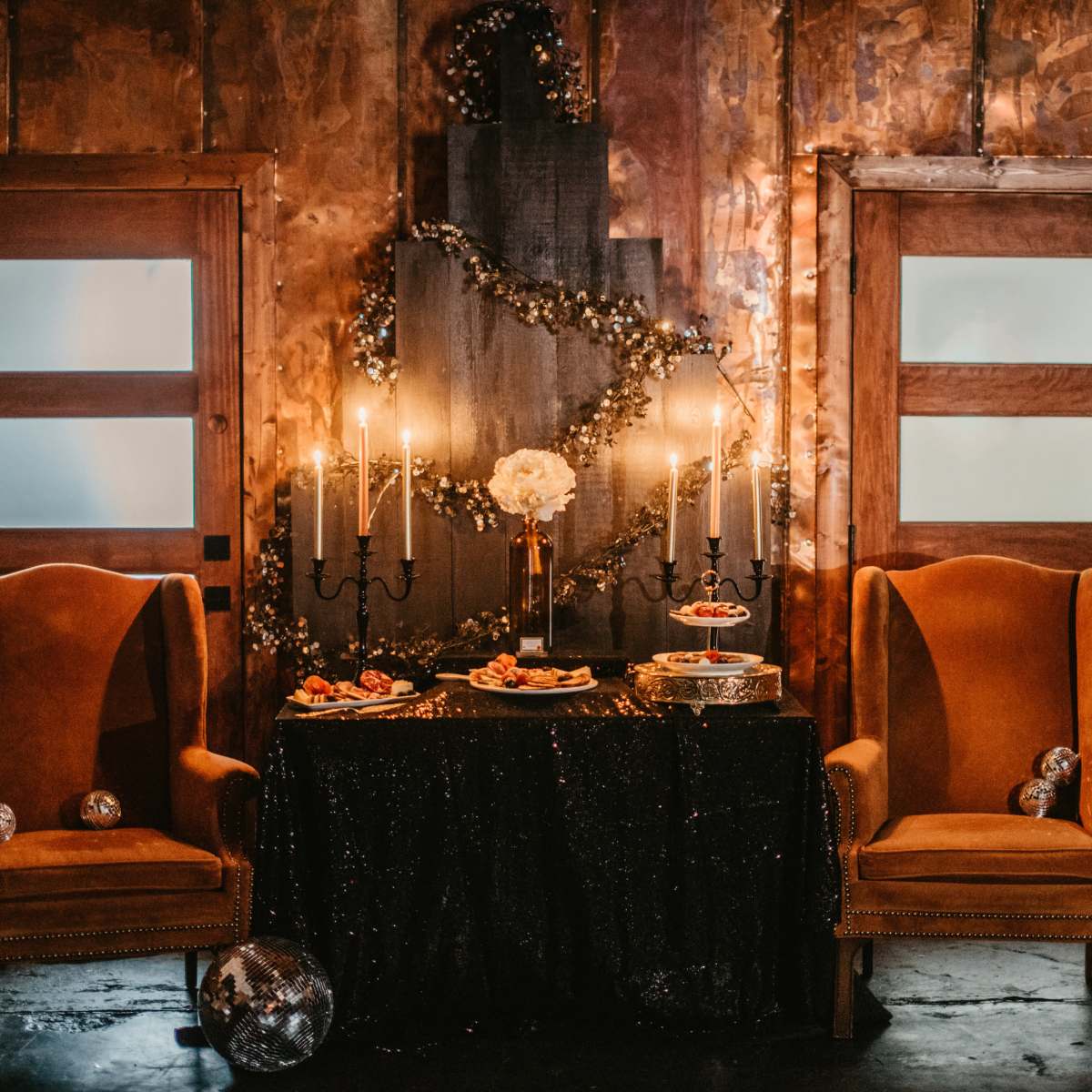The scene features two orange armchairs flanking a black table adorned with a cake and snacks, set against a backdrop of wooden walls decorated with garlands and candles.