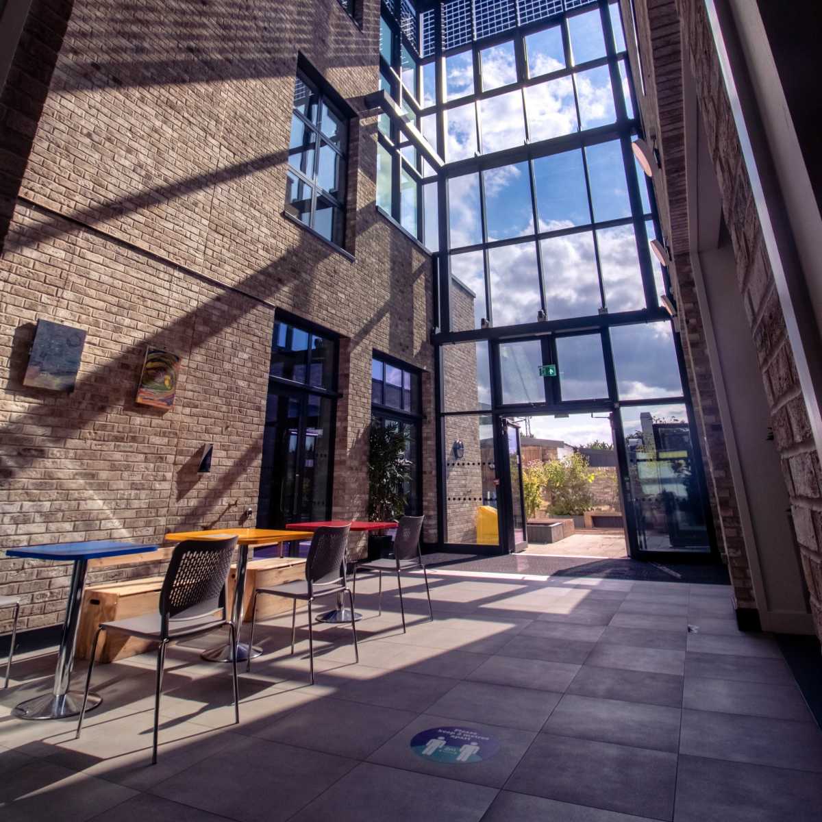 A modern architectural space with large windows, exposed brick walls, and a series of tables and chairs arranged along the floor.