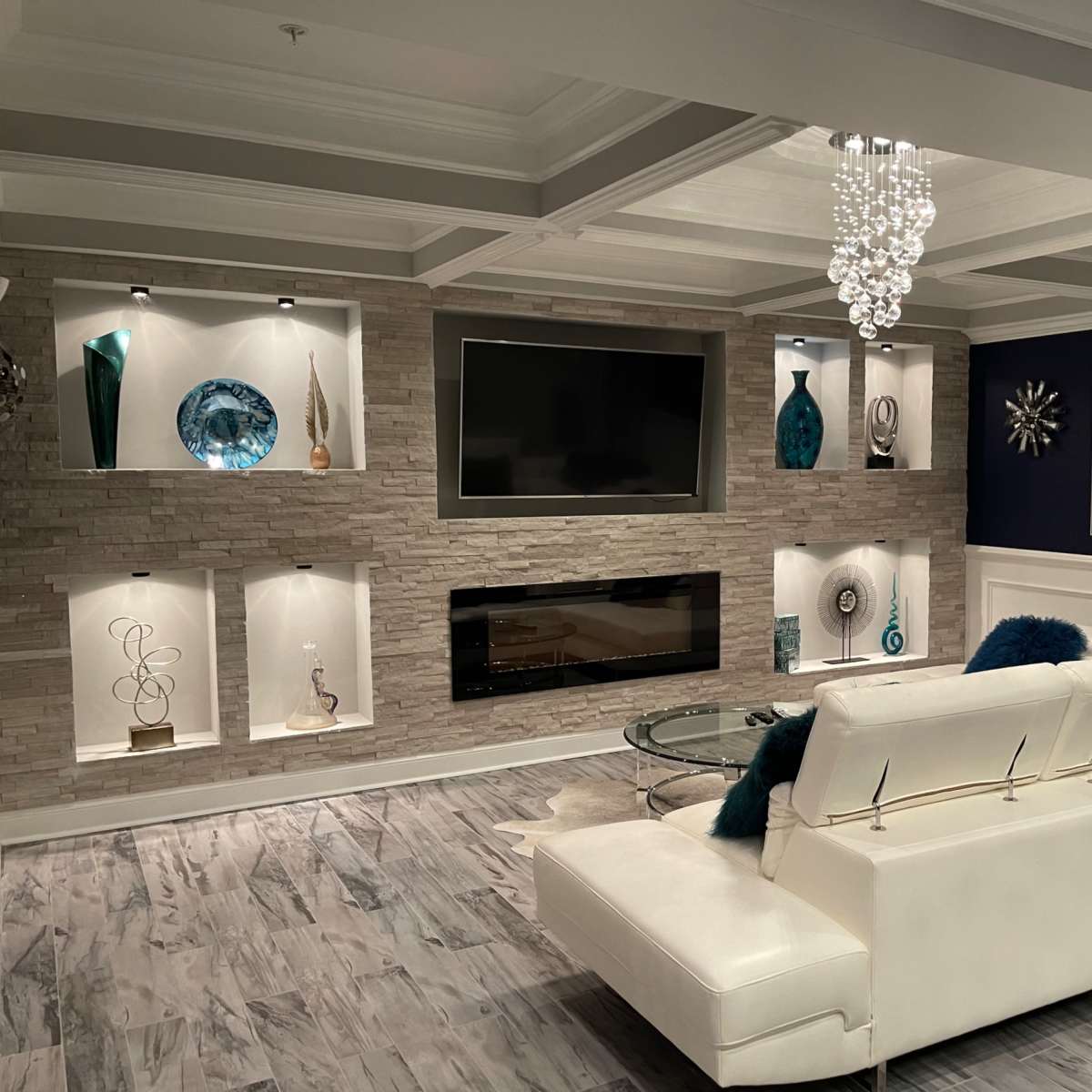 A modern living room featuring a white sectional sofa, a wall-mounted television, a stone accent wall with decorative shelves, and a glass coffee table under a chandelier.