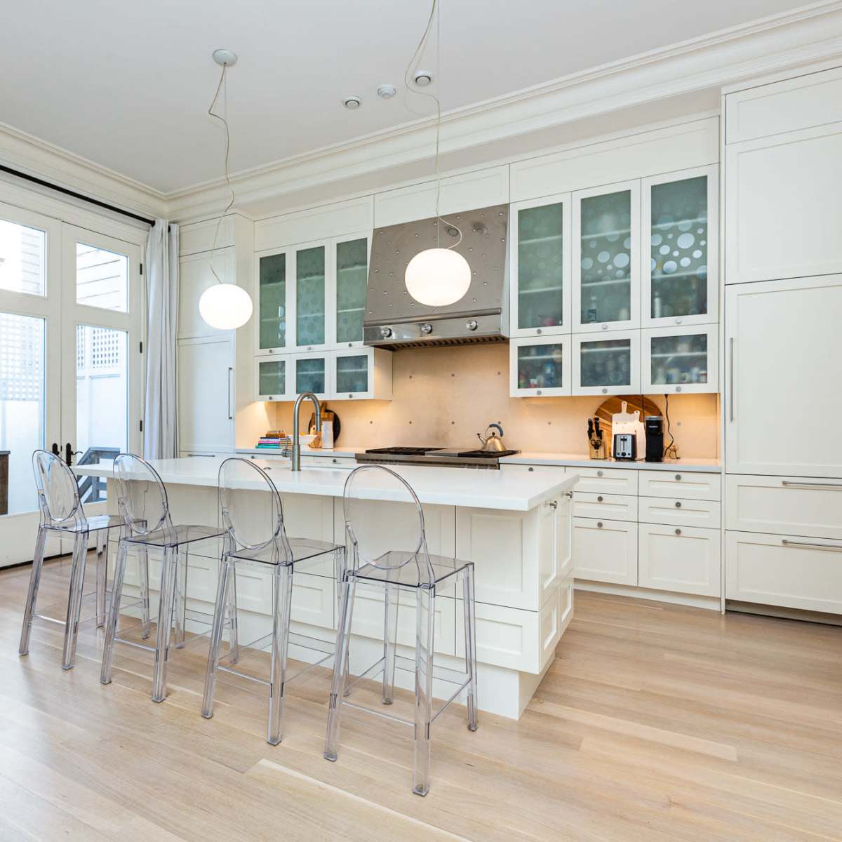 A modern kitchen features a central island with transparent stools, white cabinetry, and large windows that provide natural light.