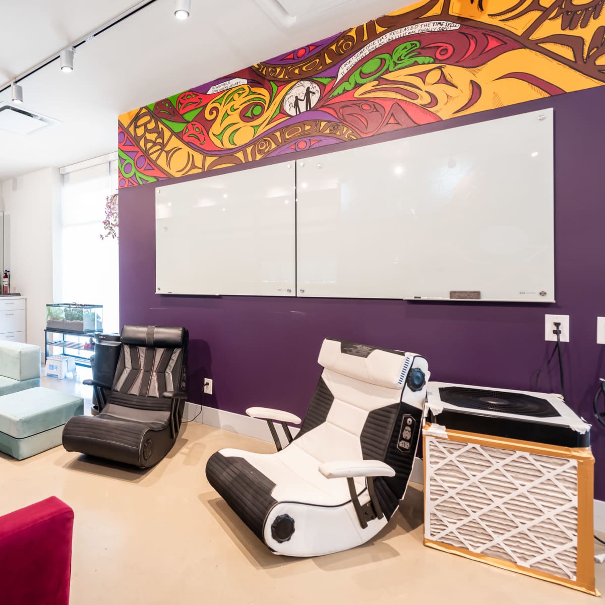A modern lounge area with colorful walls, seating options including a red velvet couch and two lounge chairs, and a kitchenette in the background.
