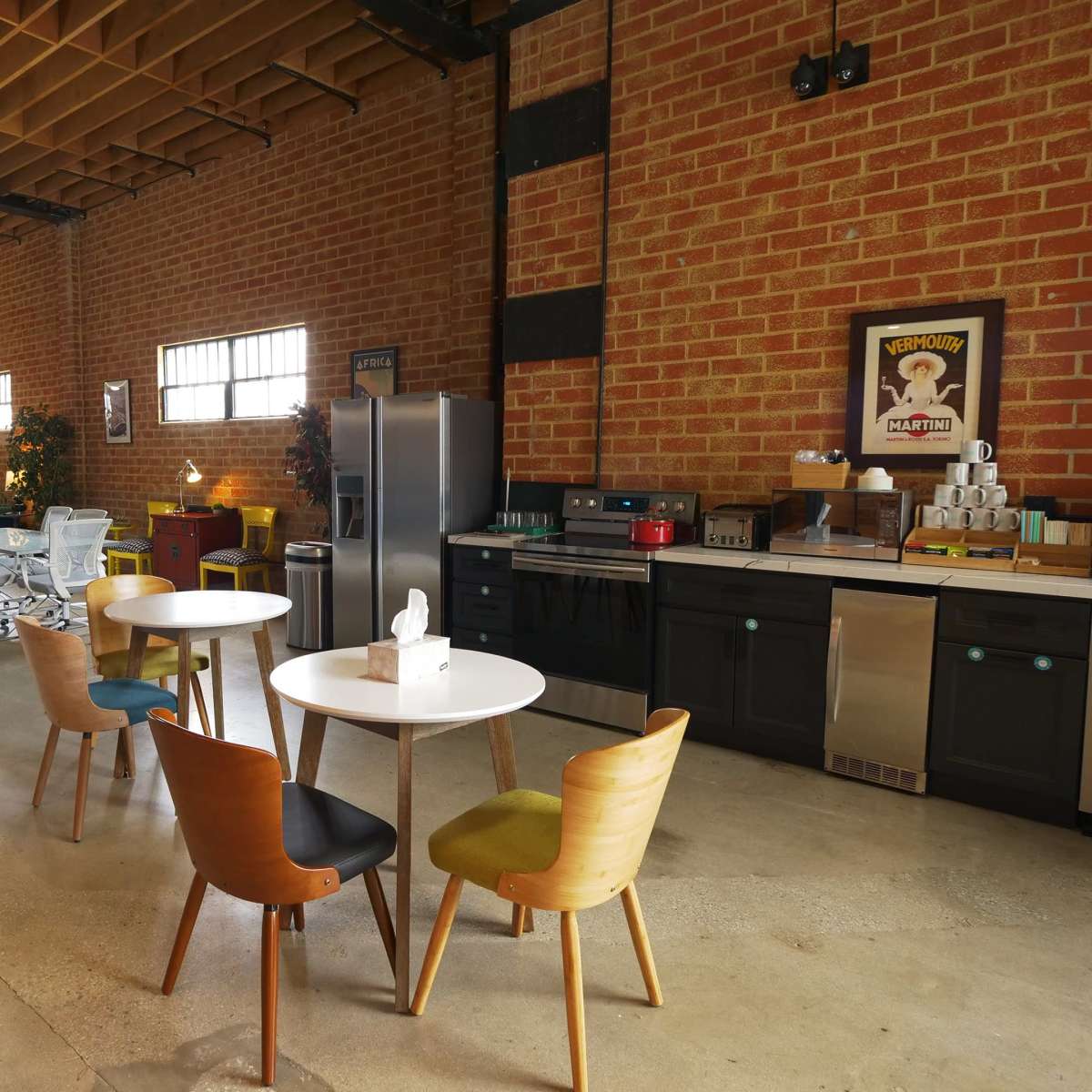 A spacious industrial-style kitchen with a dining area featuring colorful chairs around a white table, alongside modern appliances and brick walls.