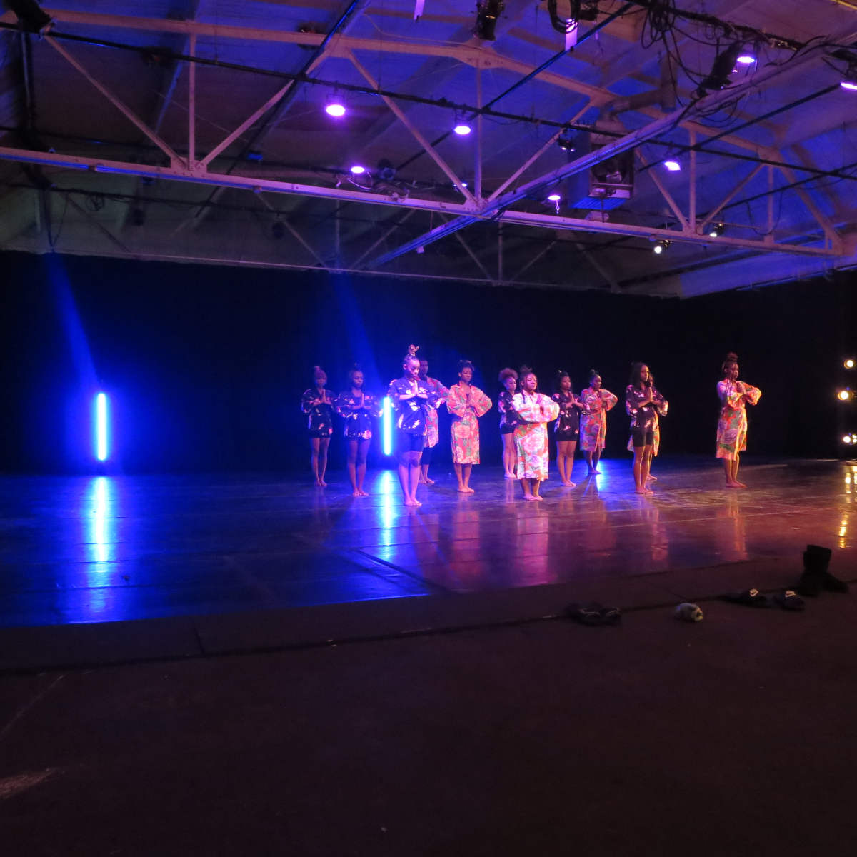 A group of performers dressed in colorful outfits is dancing on a stage illuminated by purple lights.