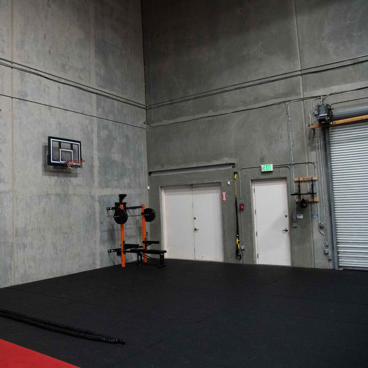 A concrete gym interior with a basketball hoop mounted on the wall, a barbell set on a rack, and a section of rubber flooring.