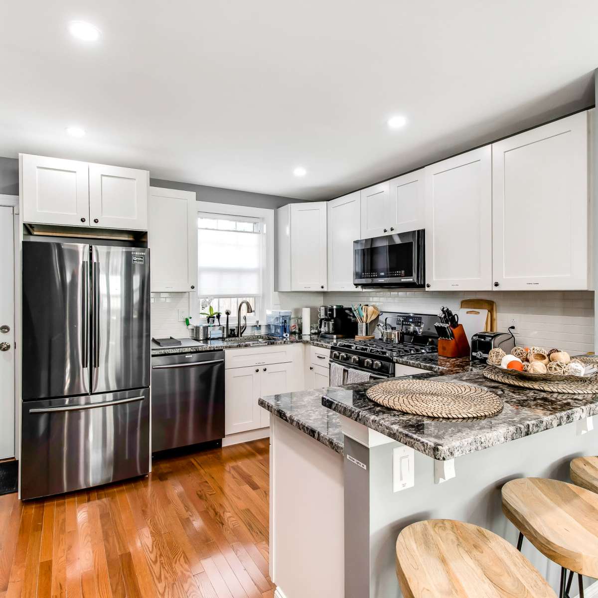 A modern kitchen features white cabinets, stainless steel appliances, and a granite countertop with wooden stools adjacent to it.