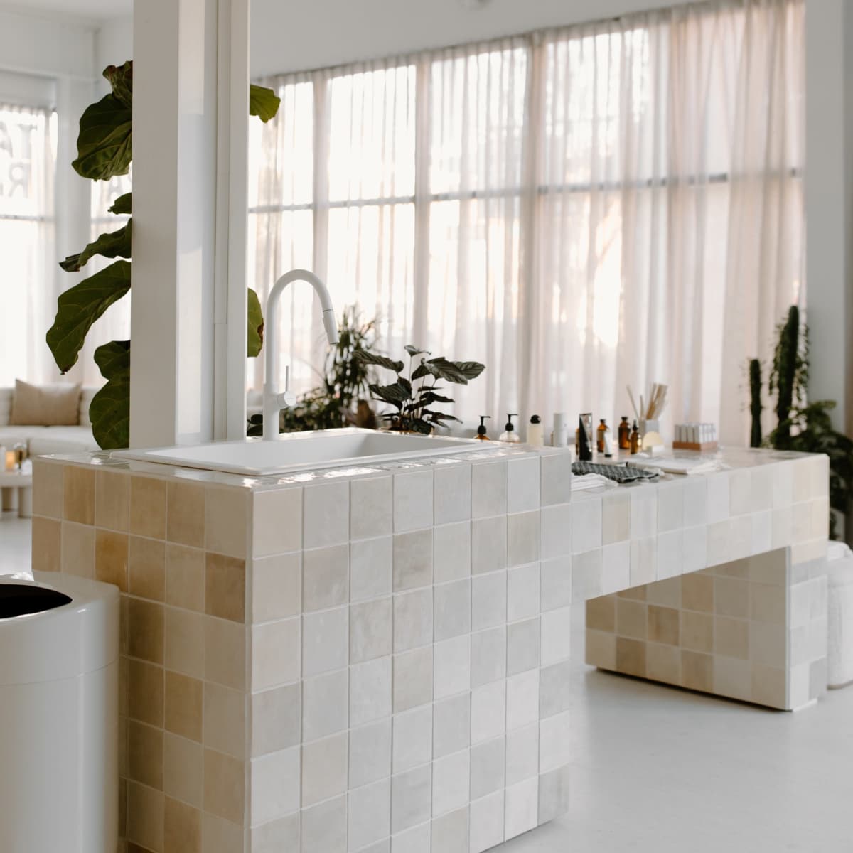 A modern, light-filled interior features a tiled counter and sink, surrounded by plants and a white trash bin.