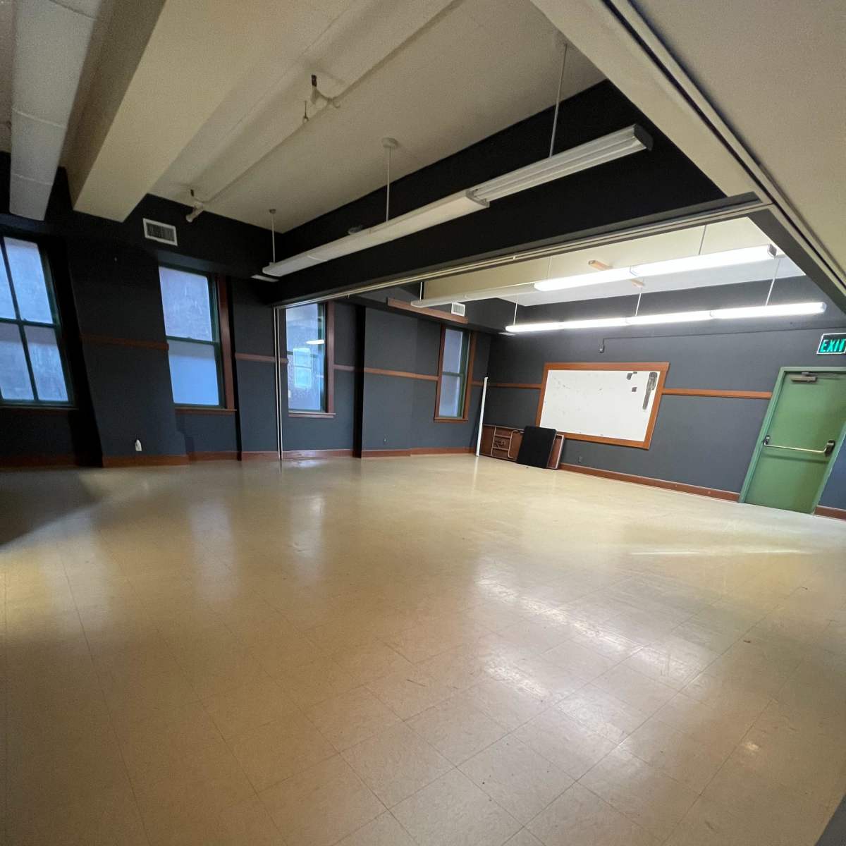 A spacious, empty room with wooden floors, large windows, and a chalkboard on the wall.