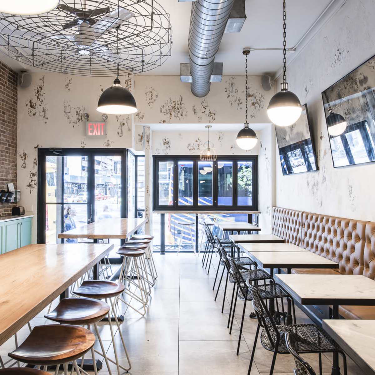 A modern café interior with wooden and metal tables, brown tufted seating, and large windows allowing natural light.