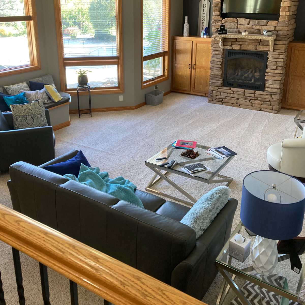 A spacious living room features a stone fireplace, two seating areas with various cushions, a coffee table with books, and large windows allowing natural light to enter.