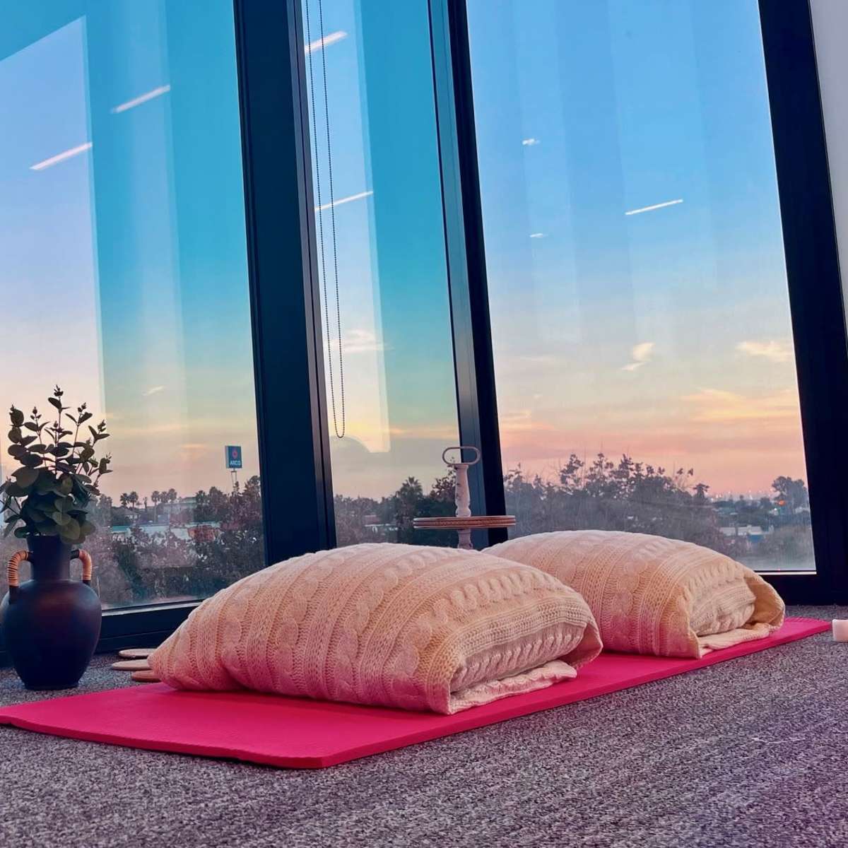 A serene room with two large cushions on a pink mat, placed beside a potted plant, all framed by floor-to-ceiling windows that overlook a colorful sunset.