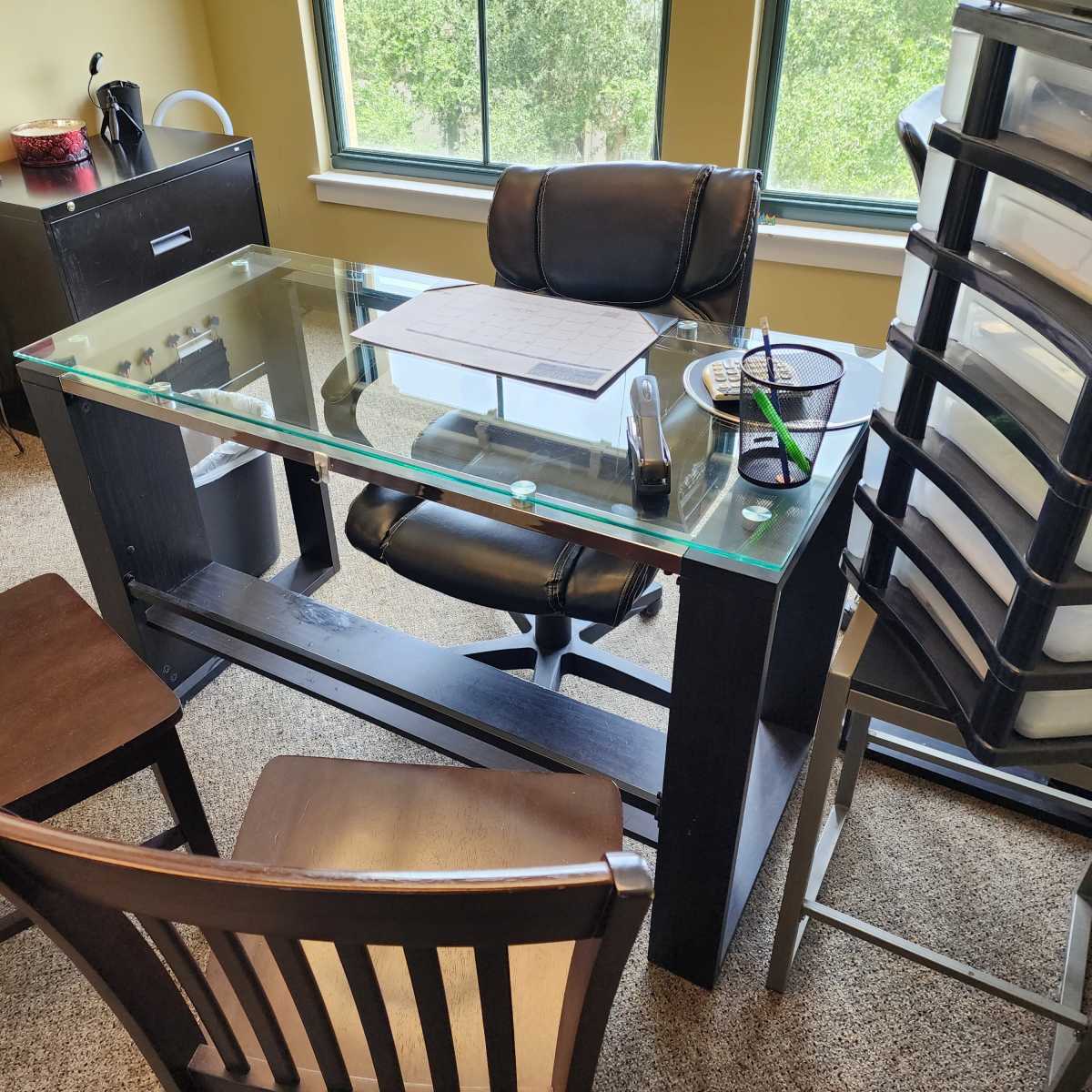 An office scene features a glass-top desk with a chair and two wooden chairs arranged around it, along with storage containers and a window revealing greenery outside.