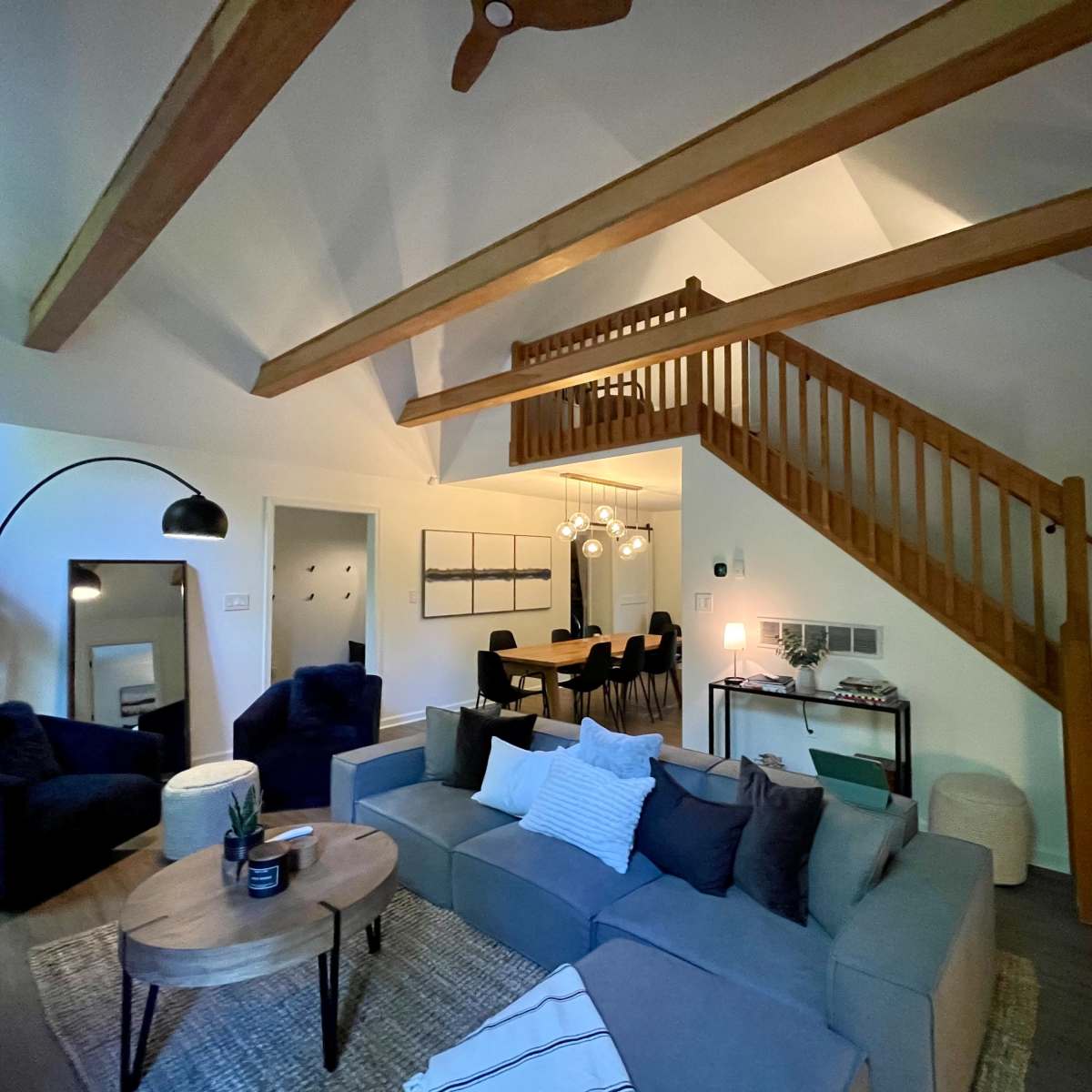 A spacious living area with a high ceiling, wooden beams, a large sectional sofa, a dining area in the background, and a staircase leading to an upper level.